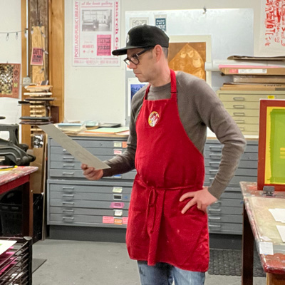 Robert Brochu looking at a silkscreen print wearing a red apron at Pickwick Independent Press in Portland, Maine